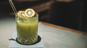 Ein erfrischendes Getränk in einem Glastumbler mit Kiwi-Scheiben und einem Strohhalm. Das Glas steht auf einem Untersetzer auf einem Tisch.
