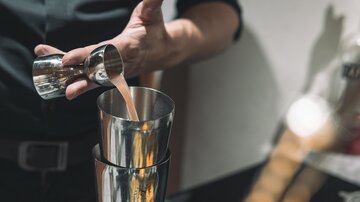 Ein Barkeeper gießt einen Cocktail in einen Shaker. Die Szene zeigt die Zubereitung eines Getränks mit glänzendem Edelstahl.