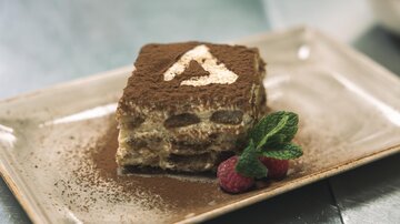 Ein köstliches Stück Tiramisu auf einem Teller, garniert mit frischen Himbeeren und einem Minzblatt. Das Dessert ist mit Kakaopulver bestäubt und hat eine dekorative Verzierung.