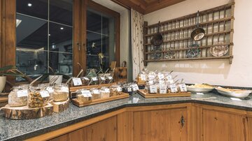 Ein charmanter Frühstücksbereich mit verschiedenen Wurst- und Käsetabletts, ergänzt durch Gläser mit selbstgemachten Marmeladen. Rundum ist eine gemütliche Holzverkleidung zu sehen.