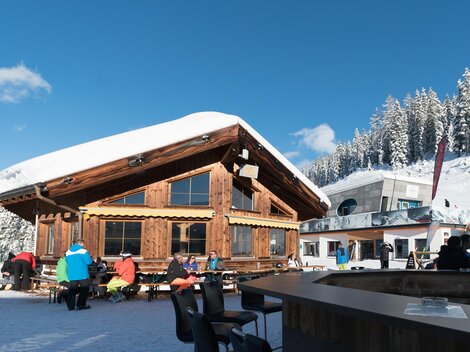 Eine gemütliche Berghütte im Schnee mit Gästen im Freien. Im Hintergrund sind schneebedeckte Berge und blauer Himmel zu sehen.