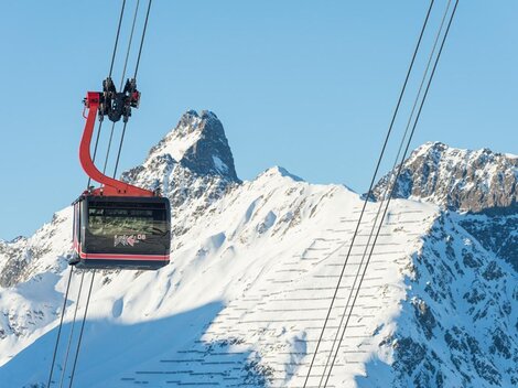 A cable car glides over snow-covered mountains. The sky is clear and blue.