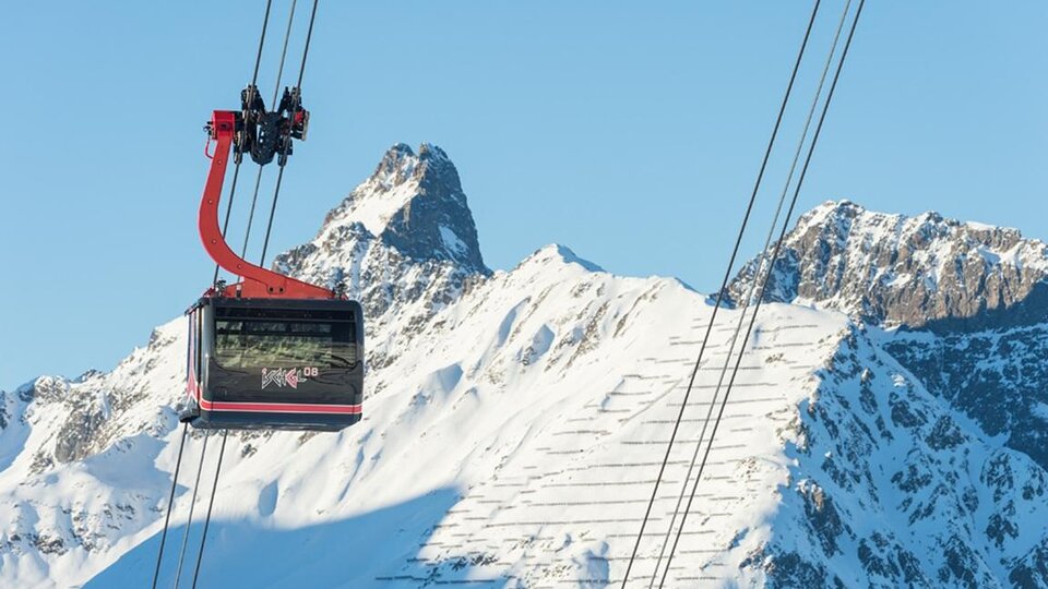 A cable car glides over snow-covered mountains. The sky is clear and blue.