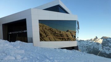 A modern building in the mountains, surrounded by snow. The large glass front reflects the impressive mountain landscape.