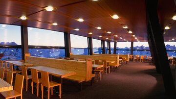 A modern restaurant with large windows and a view of the mountains. The decor is simple with wooden tables and chairs.