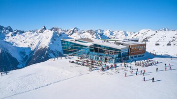 A modern mountain restaurant in the Alps surrounded by snow-covered mountains. Skiers enjoy the sunny winter landscape.