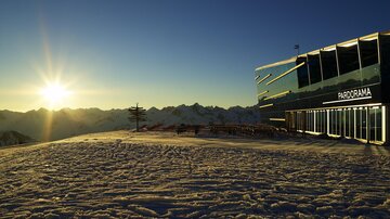 A modern building stands in a snow-covered landscape. In the background, the mountains and the sun can be seen illuminating the sky.
