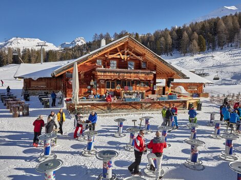 A cozy mountain cabin in the snowy landscape, surrounded by trees. Many people enjoy the winter atmosphere and stand at tables outdoors.