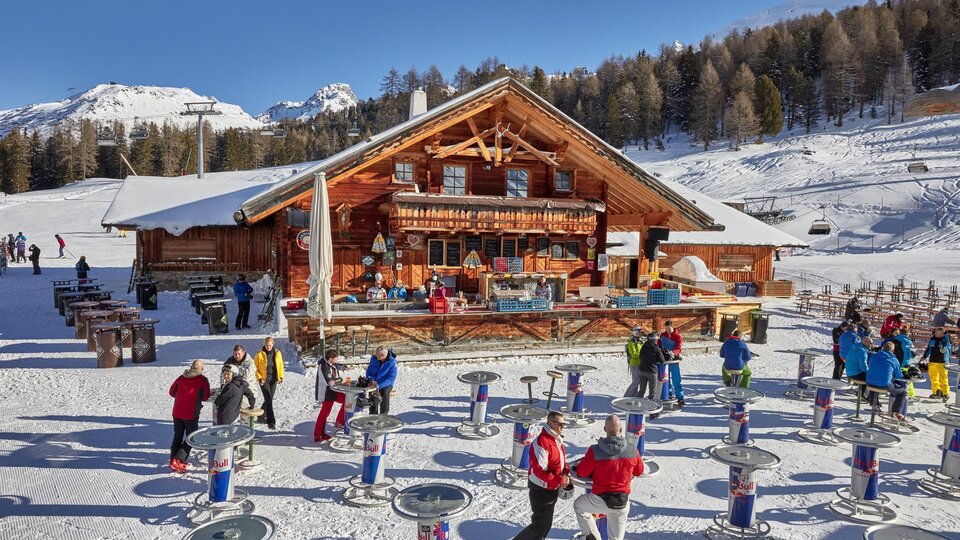 Eine gemütliche Berghütte in der verschneiten Landschaft, umgeben von Bäumen. Viele Menschen genießen die winterliche Atmosphäre und stehen an Tischen im Freien.