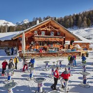 Eine gemütliche Berghütte in der verschneiten Landschaft, umgeben von Bäumen. Viele Menschen genießen die winterliche Atmosphäre und stehen an Tischen im Freien.