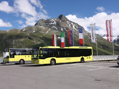 Zwei gelbe Busse stehen in einer Parkbucht vor einer beeindruckenden Berglandschaft. Im Hintergrund wehen Flaggen verschiedener Länder im klaren Himmel.