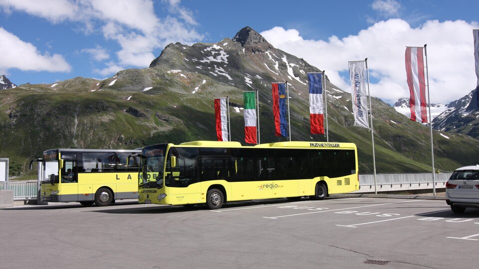 Zwei gelbe Busse stehen in einer Parkbucht vor einer beeindruckenden Berglandschaft. Im Hintergrund wehen Flaggen verschiedener Länder im klaren Himmel.