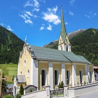 Eine malerische Kirche mit einem hohen Turm und einer gelben Fassade, umgeben von grünen Bergen. Der klare Himmel lässt die Landschaft strahlend wirken.