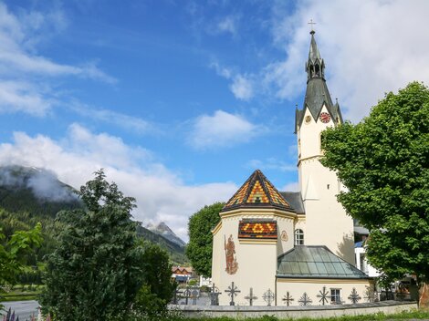 A picturesque church with a tall tower and a colorful roof. In the background, there are green mountains and a blue sky.