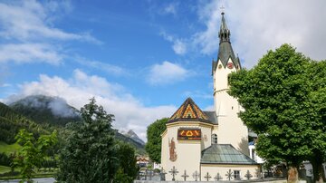 A picturesque church with a tall tower and a colorful roof. In the background, there are green mountains and a blue sky.