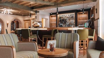 A stylishly furnished café with comfortable armchairs and a central table. In the background, a well-stocked bar is visible.