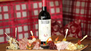 Ein schöner Teller mit verschiedenen Wurst- und Käseaufschnitten, dekoriert mit Gemüse und Oliven. Dazu steht eine Flasche rotwein auf einem Tisch in einem rustikalen Ambiente.
