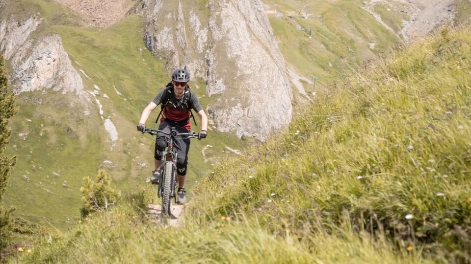 A cyclist rides on a narrow path through the green mountainous landscape. In the background, rocky mountains and a clear sky can be seen.