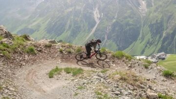 A cyclist rides on a winding path in the mountains. In the background, green meadows and majestic mountain peaks can be seen.