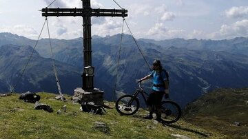 A person stands with their bicycle at a summit cross in the mountains. In the background, green hills and a clear blue sky are visible.