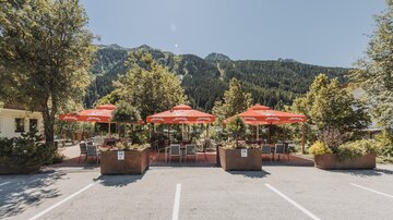 An inviting café with red umbrellas, surrounded by trees. In the background, shady mountains can be seen.