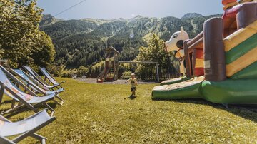A beautiful outdoor playground with a large bounce house and a picturesque mountain landscape in the background. In the foreground, there are deck chairs on the grass.
