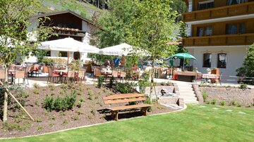 An inviting outdoor seating area with tables and chairs, surrounded by trees and beautiful greenery. In the background, a charming building can be seen.