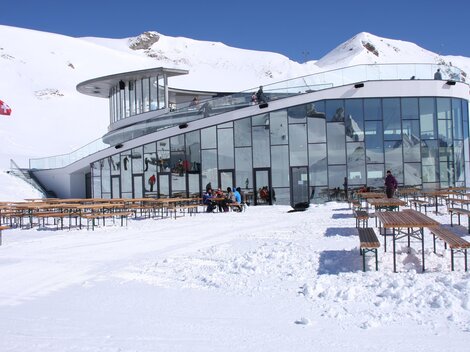 A modern building in the mountains, surrounded by snow. Outside, there are rows of wooden benches and people enjoying the wintry landscape.