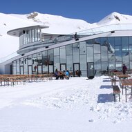 Ein modernes Gebäude in den Bergen, umgeben von Schnee. Draußen stehen Holzbankreihen und Menschen genießen die winterliche Landschaft.
