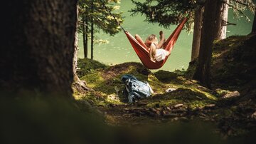 A person relaxes in a hammock between trees by the shore of a lake. Next to the hammock, there is a backpack on the moss-covered ground.