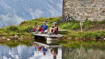 Two cyclists are sitting on a pier at a clear pond. In the background, there are mountains and an old stone wall.