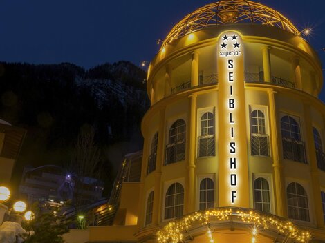 A brightly lit building named "Seiblshof" stands in the evening. In the background, snow-covered mountains are visible.