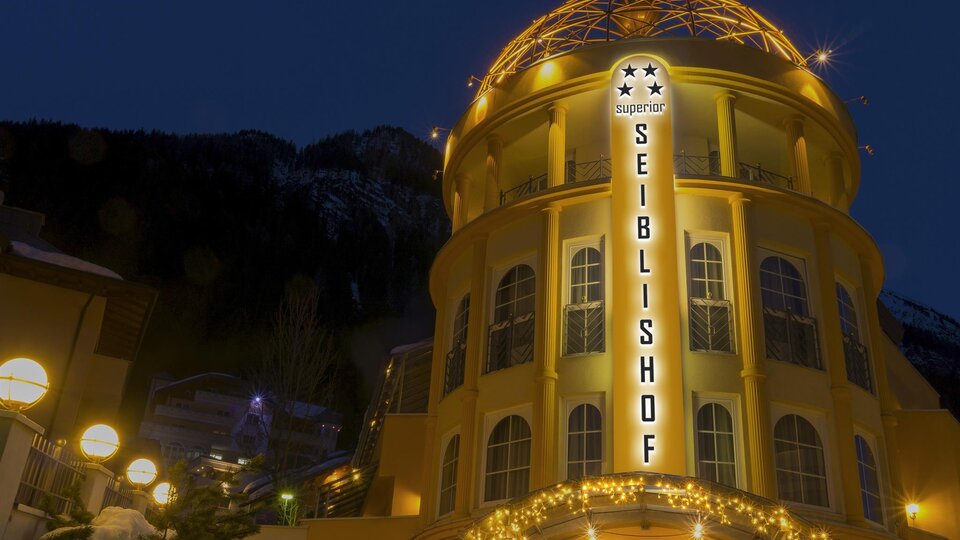 A brightly lit building named "Seiblshof" stands in the evening. In the background, snow-covered mountains are visible.