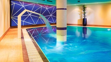 An elegant indoor swimming pool with smooth water and a modern water feature. The walls are in soft colors, and a plant adds fresh accents.
