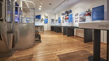 Ein moderner Ausstellungsraum mit Holzfußboden und blauen Informationswänden. Verschiedene Exponate und Tische sind im Raum angeordnet.