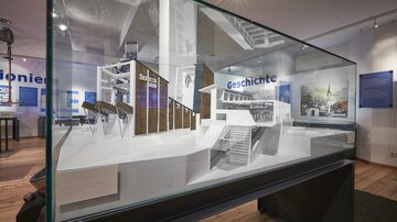 Eine Vitrine mit einem Architekturmodell, das verschiedene Gebäude zeigt. Der Hintergrund enthält informative Texte und Bilder zur Geschichte.