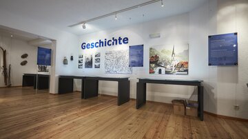 Ein moderner Raum mit Holzfußboden, der eine Ausstellung zum Thema Geschichte zeigt. An der Wand hängen erläuternde Tafeln und Bilder.