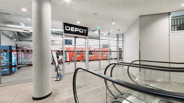 A modern entrance area with an escalator and a large "DEPOT" sign. In the background, red shelves are visible.
