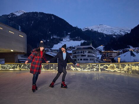A couple is ice skating under a clear evening sky. Snow-covered mountains and illuminated buildings can be seen in the background. | © Silvrettaseilbahn AG