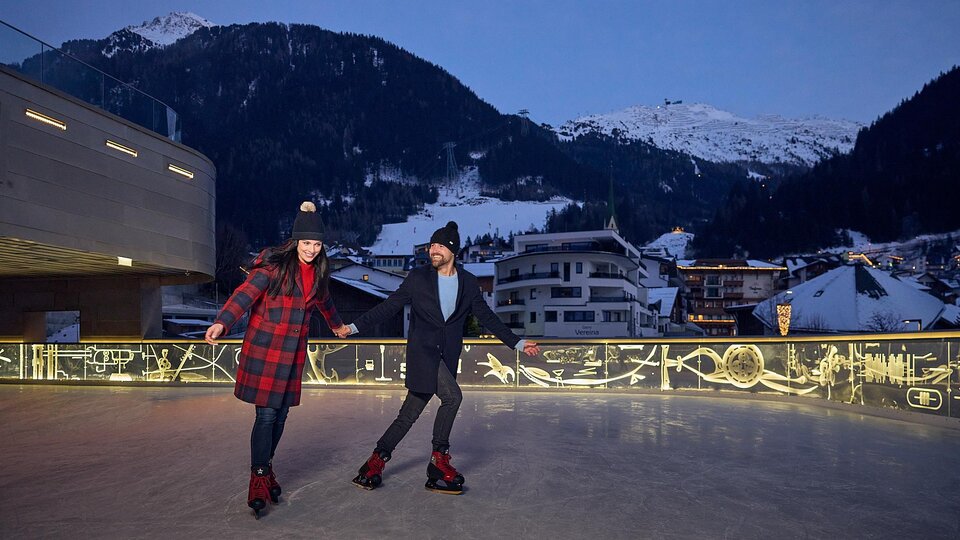 A couple is ice skating under a clear evening sky. Snow-covered mountains and illuminated buildings can be seen in the background. | © Silvrettaseilbahn AG