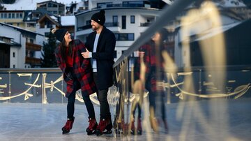 A couple is laughing and enjoying ice skating on an ice rink. In the background, snowy buildings and a wintry landscape are visible.