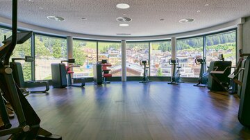 A modern fitness room with several machines and large windows. Outside is a beautiful view of a city with green hills.