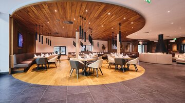 Ein modernes Restaurant mit holzverkleideter Decke und stilvollen Sitzbereichen. Die Einrichtung ist hell und einladend, mit runden Tischen und eleganten Stühlen.