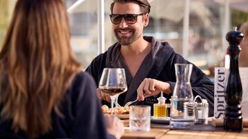 Ein Mann sitzt lächelnd an einem Tisch und genießt ein Essen. Im Hintergrund sind Gläser und Essig-Öl-Set zu sehen, während er mit einer Person spricht.