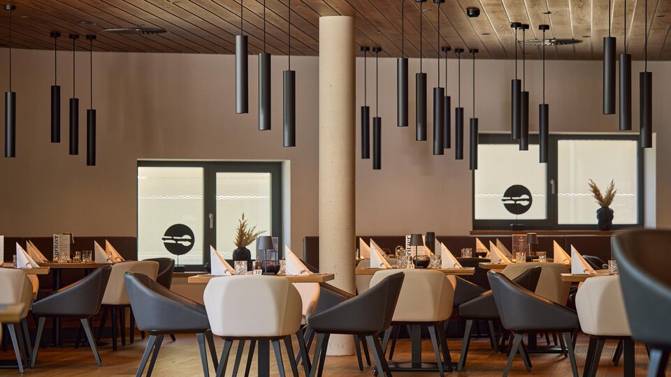 Ein modernes Restaurant mit eleganten Tischen und Stühlen. Die Decke ist mit schlichten Hängeleuchten dekoriert und große Fenster sorgen für viel Licht.