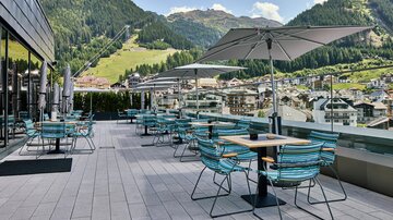Eine gemütliche Terasse mit blauen Stühlen und Tischen, umgeben von Bergen. Sonnenschirme bieten Schatten und der Blick auf die Stadt ist atemberaubend.
