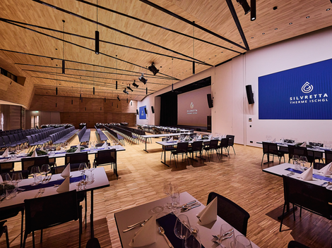 A modern event room with several tables elegantly set. The room has a large screen and wooden paneling on the walls.