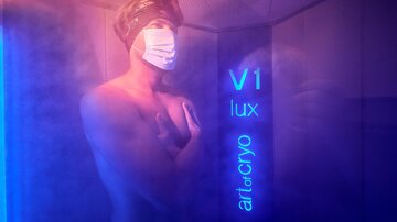 A man stands in a cool room with blue lighting and wears a facial mask. He looks relaxed and is using a cryotherapy treatment.