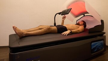 A man lies relaxed on a lounge under a special treatment unit. Red light beams are directed at him as he stretches out a hand.