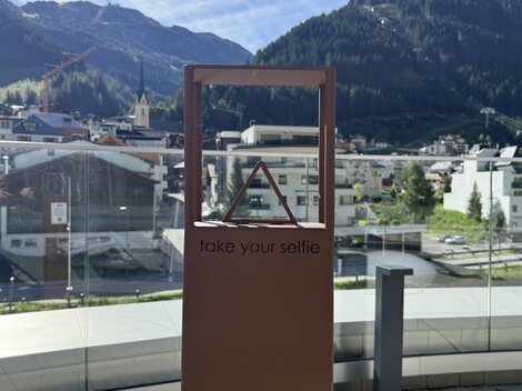 Ein Selbstporträt-Rahmen mit der Aufforderung "take your selfie" steht vor einer Berglandschaft. Im Hintergrund sind Gebäude und grüne Flächen sichtbar.
